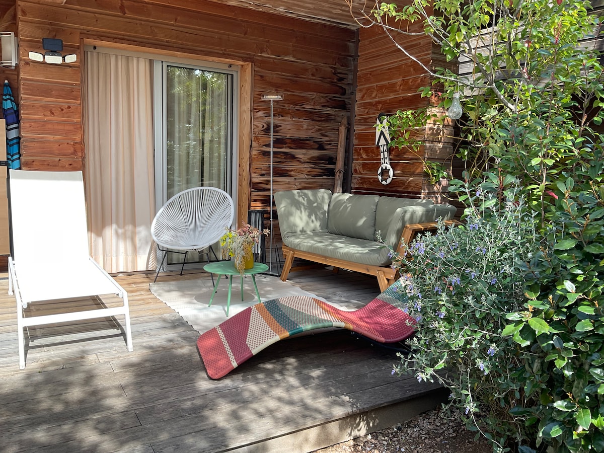 An outdoor seating area is presented, featuring a comfortable green sofa, a round table, and a lounge chair. Lush greenery and flowering plants surround the space, while light wood paneling complements the natural setting. Soft sunlight filters through sheer curtains by the entrance.