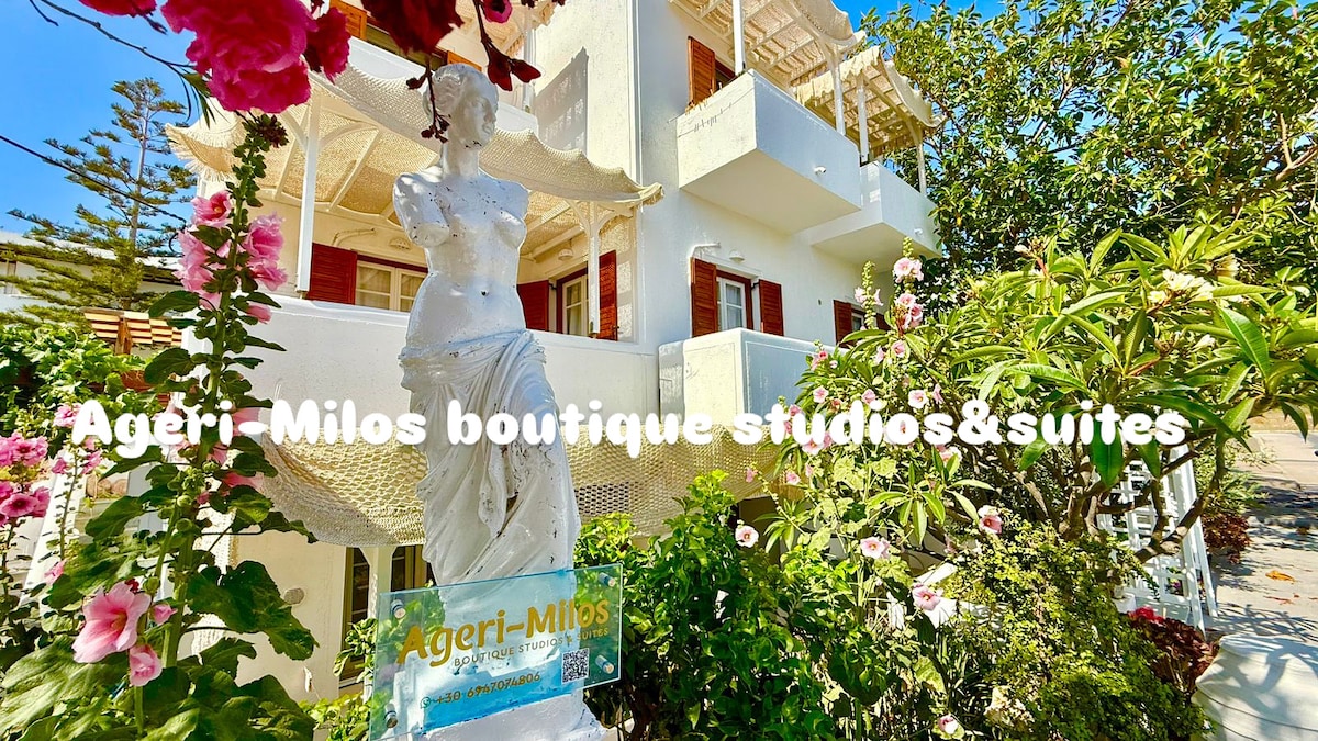 The exterior of the boutique studio is surrounded by vibrant greenery and blooming flowers. A statue resembling a classical figure stands prominently at the entrance, while a welcoming sign indicates the property name, blending with the white-washed architecture of the building.