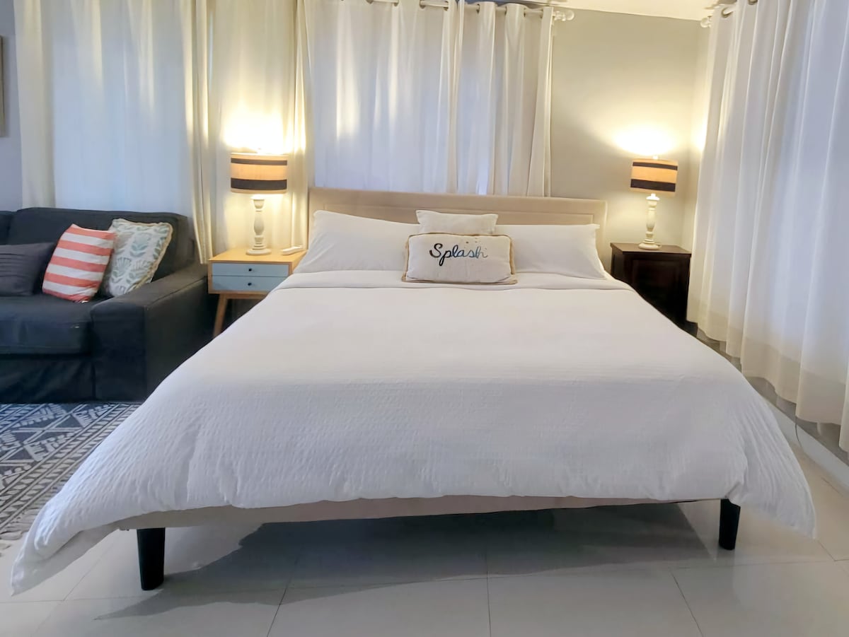 A spacious sleeping area features a king-size bed with a white duvet and decorative pillow. Soft lighting is provided by bedside lamps, with sheer white curtains allowing natural light to filter through. A cozy seating area is visible in the background.