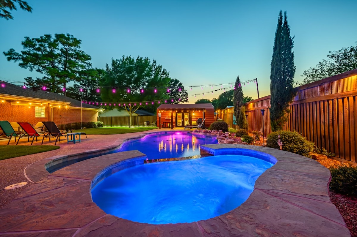 A serene backyard oasis features a glistening pool and hot tub, both illuminated in soft blue. String lights enhance the atmosphere above carefully arranged seating. Lush greenery borders the space, creating a tranquil environment for relaxation.