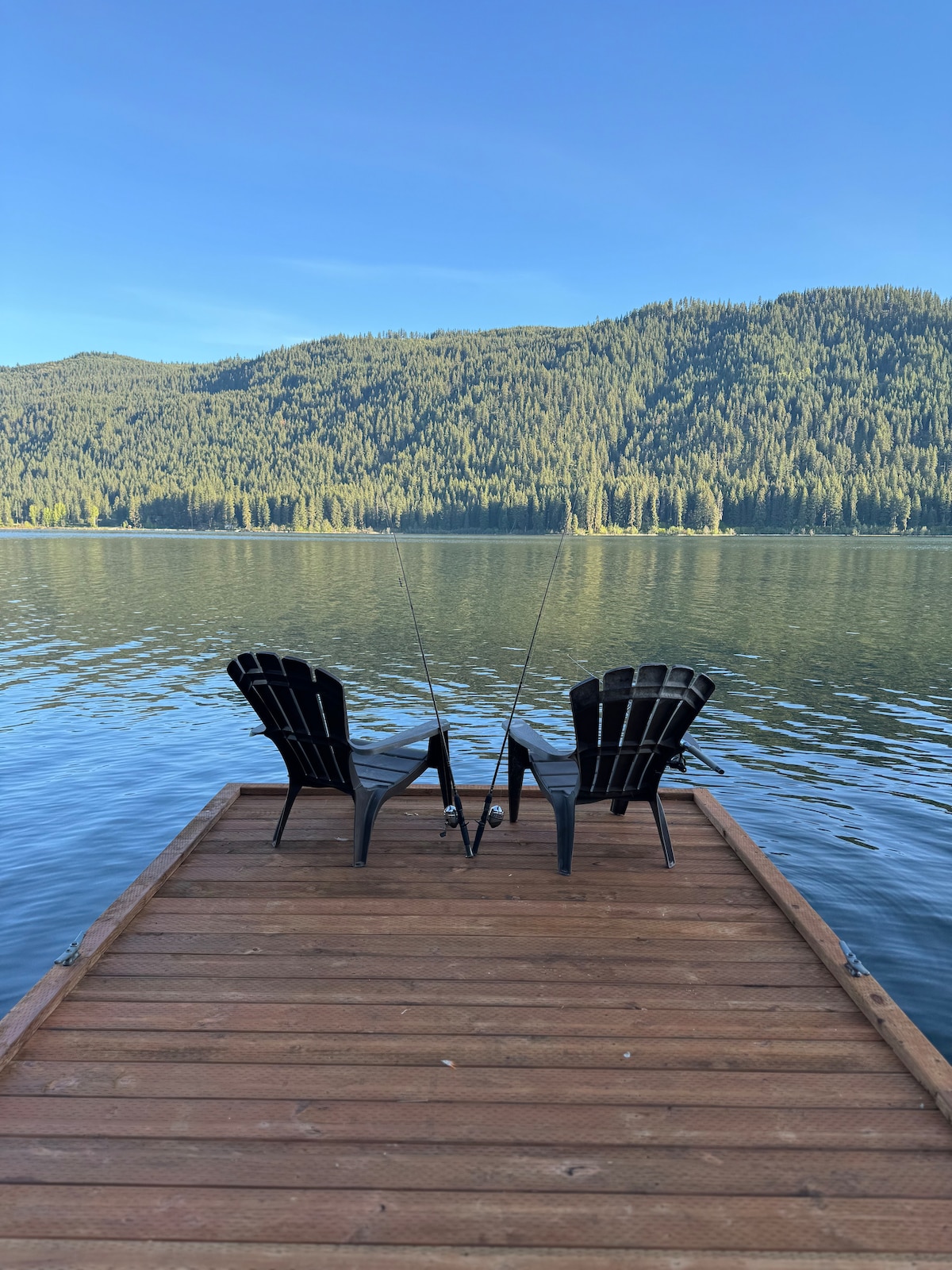 A wooden dock extends over calm waters, providing a serene spot for fishing. Two black Adirondack chairs are positioned to face the lake, complemented by fishing rods resting between them. Lush green hills rise in the background, reflecting in the tranquil surface.