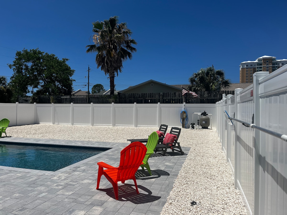 A spacious outdoor area is shown with a swimming pool and surrounding lounge chairs in vibrant colors. A palm tree is visible against a clear blue sky, and a white privacy fence borders the perimeter, providing a sense of seclusion. Gravel is laid out around the pool deck.