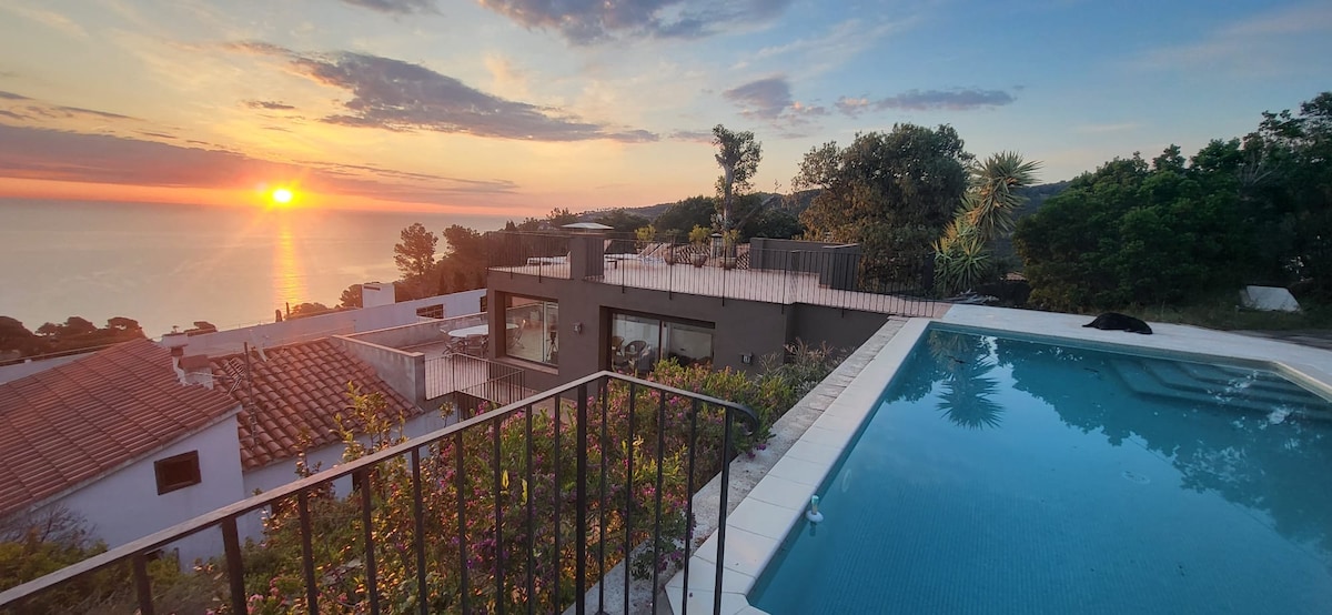 A private rooftop pool reflects the sunset over the sea, with panoramic views of the surrounding hills and greenery. The terrace is adorned with plants, complementing the serene atmosphere. The house is partially visible, showcasing modern architecture in a tranquil setting.
