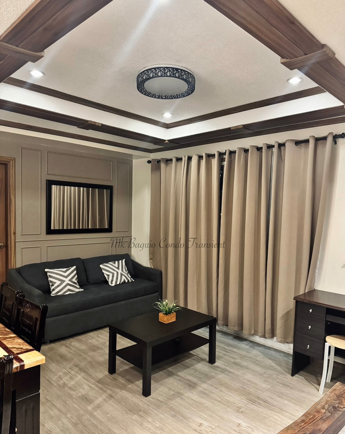 A comfortable seating area features a dark sofa adorned with patterned cushions. A small coffee table is positioned in front of the sofa, complemented by a potted plant. Soft lighting is provided by a circular ceiling fixture, while beige drapes frame the window.