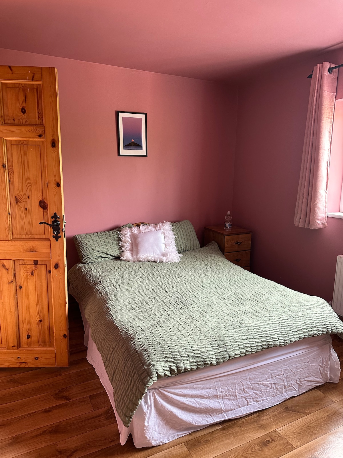 A cozy bedroom features a spacious bed covered with a light green blanket and adorned with a decorative pillow. A wooden nightstand holds a lamp, and a framed picture is displayed on the pink wall. Natural light illuminates the room through a window with soft curtain drapes.