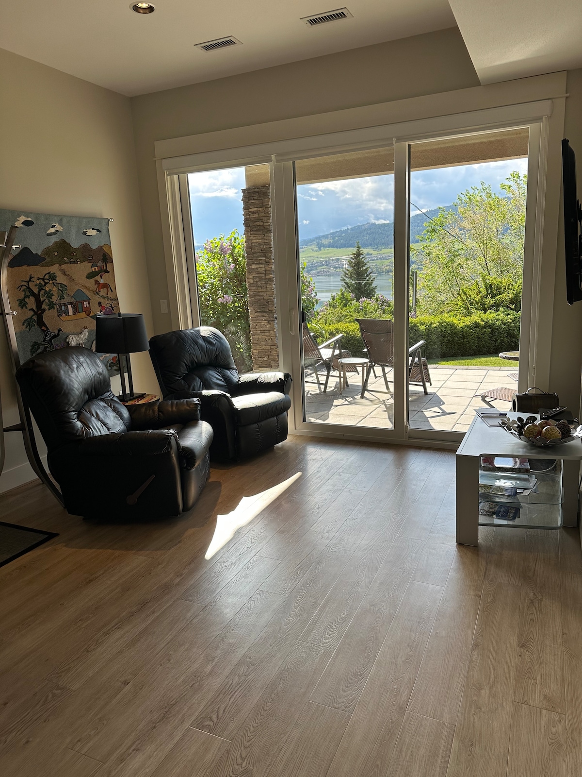 A light-filled living area features two reclining chairs set on a wooden floor. Large sliding glass doors provide a view of the garden and patio area, where a dining set is visible amidst greenery and the surrounding hills.