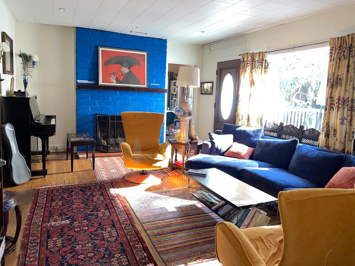 A cozy living room is featured, highlighted by a vibrant blue accent wall. Modern seating includes two yellow chairs and a plush sectional sofa in deep blue, surrounding a coffee table. A wooden piano and a colorful rug add warmth, with large windows inviting natural light.