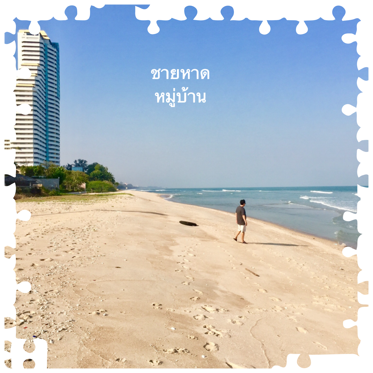 The image showcases a wide sandy beach with gentle waves lapping at the shore. A lone figure walks along the beach, leaving footprints in the soft sand. In the background, a tall building stands against a clear blue sky, providing a sense of nearby amenities.