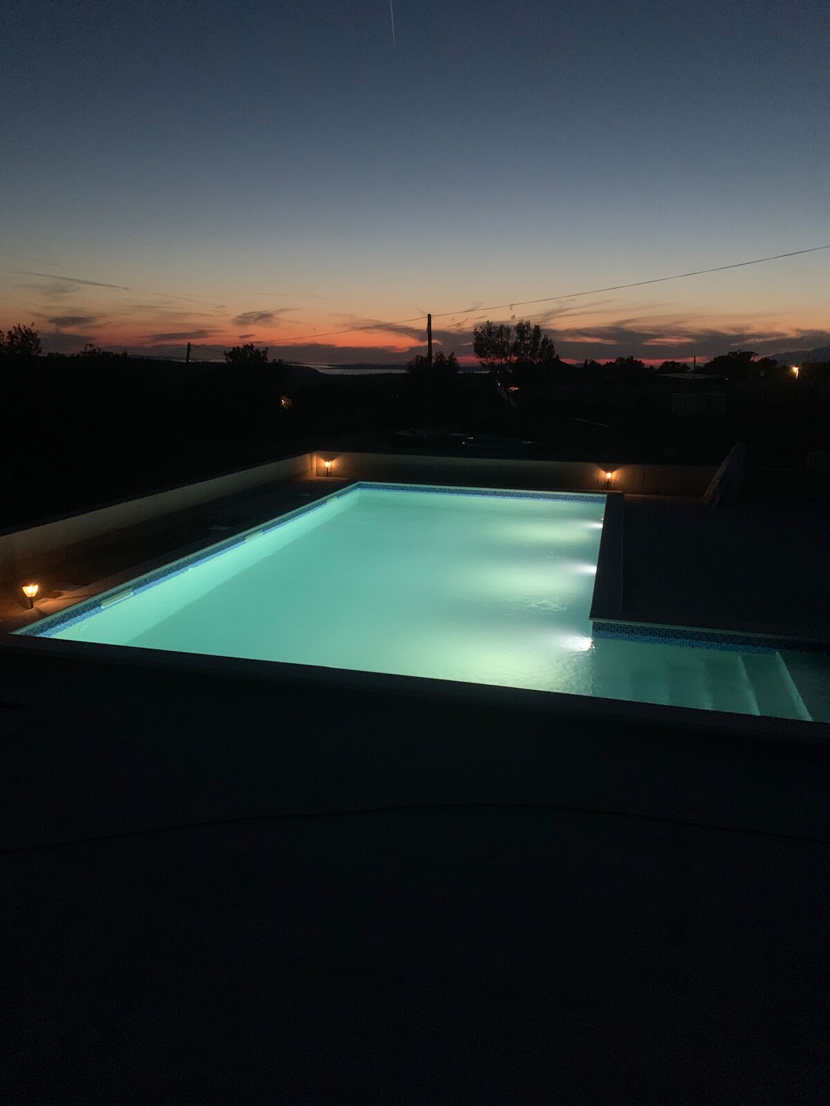 A tranquil swimming pool is illuminated by soft lighting, reflecting the calm water. The horizon displays a gradient of colors from orange to deep blue as the sun sets, with silhouettes of trees and distant structures subtly visible.