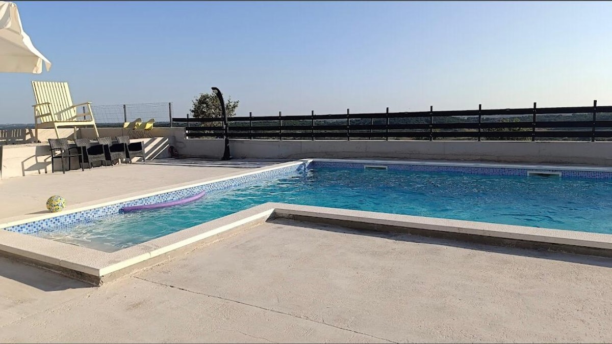 A rooftop pool area features a clear blue pool surrounded by a spacious deck. Lounge chairs are positioned near the poolside, with a shaded area provided by an outdoor umbrella. Views of the surrounding landscape are visible in the background under a clear sky.