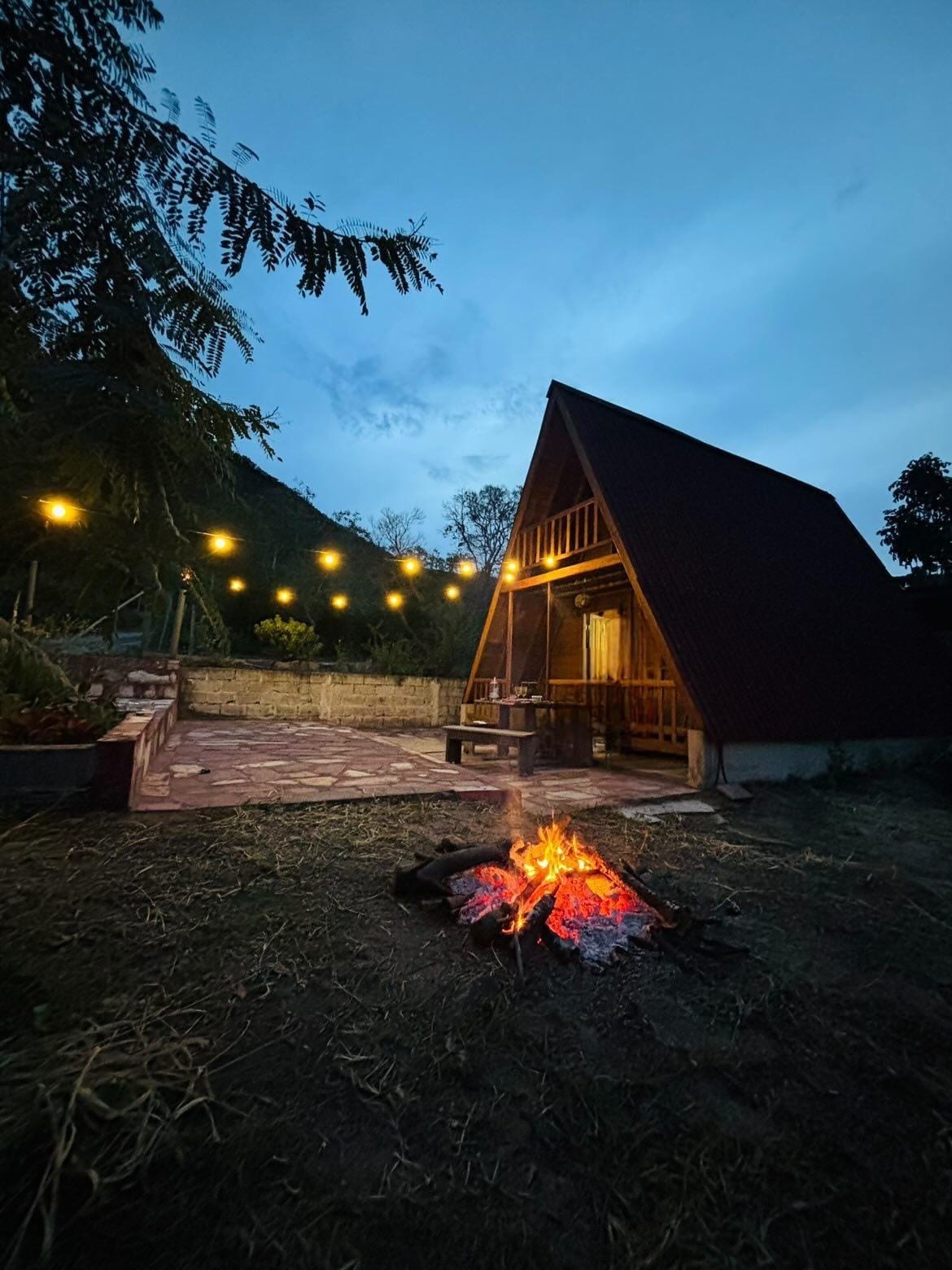 A charming duplex chalet is illuminated by warm outdoor lights as night falls. A cozy fire pit, surrounded by stones, adds a focal point to the grassy area. The A-frame design showcases wooden elements, blending harmoniously with the natural surroundings.