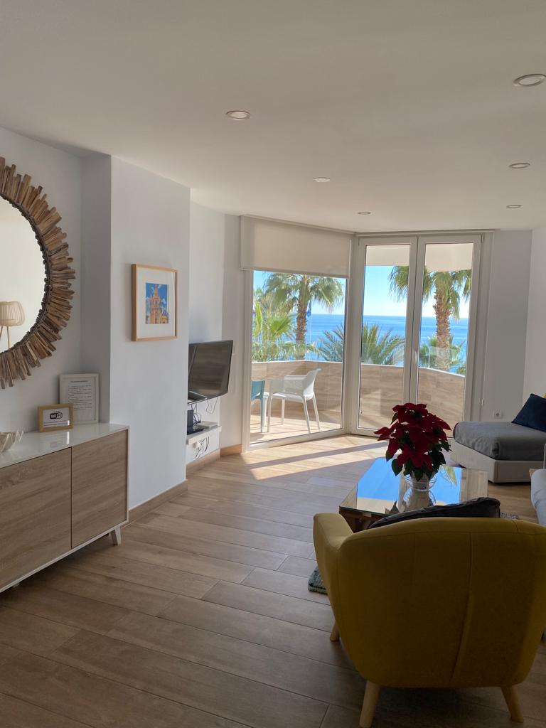 A bright living area features a circular mirror mounted on a white wall, reflecting natural light. A comfortable yellow chair is positioned beside a wooden cabinet housing a television. The large glass doors lead to a terrace with views of the beach, framed by palm trees.