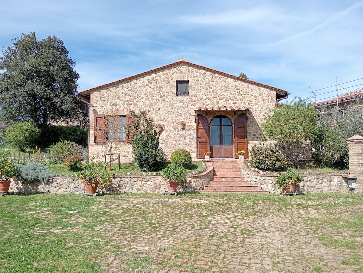 A charming stone house is framed by well-kept gardens and trees. Wooden shutters adorn the windows, while a staircase leads to the entrance. Potted plants add greenery, and a grassy area surrounds the property, providing a serene outdoor space.