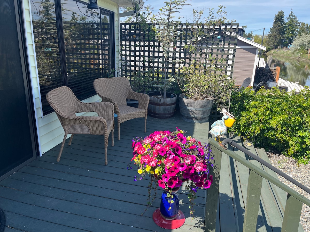 A cozy deck area features two wicker chairs positioned beside a vibrant flower arrangement in a blue vase. A garden trellis can be seen in the background, with potted plants and a colorful decorative item accenting the space. A peaceful view of the surrounding greenery is visible.