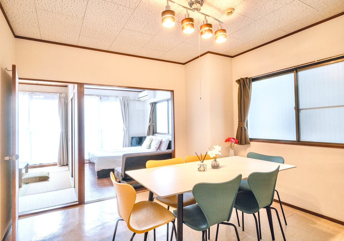 A bright dining area is displayed, featuring a rectangular table with six multi-colored chairs. Nearby, large windows allow natural light to fill the space. A cozy living area is visible through an open doorway, showcasing comfortable seating and additional windows.