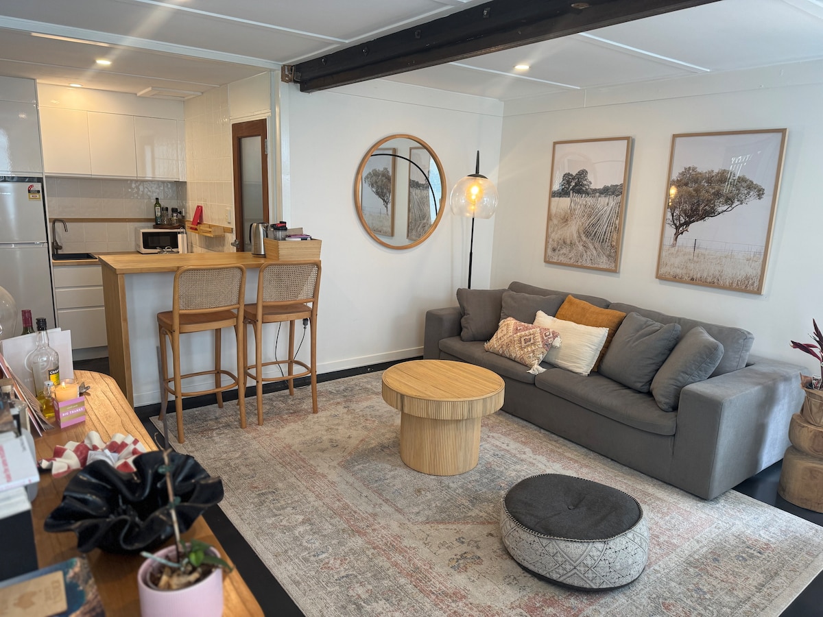 A modern living area is presented with soft gray couches and a round wooden coffee table. Two framed artworks adorn the walls, complemented by a plush accent rug. A breakfast bar with stools is visible, alongside a kitchen space designed for convenience.