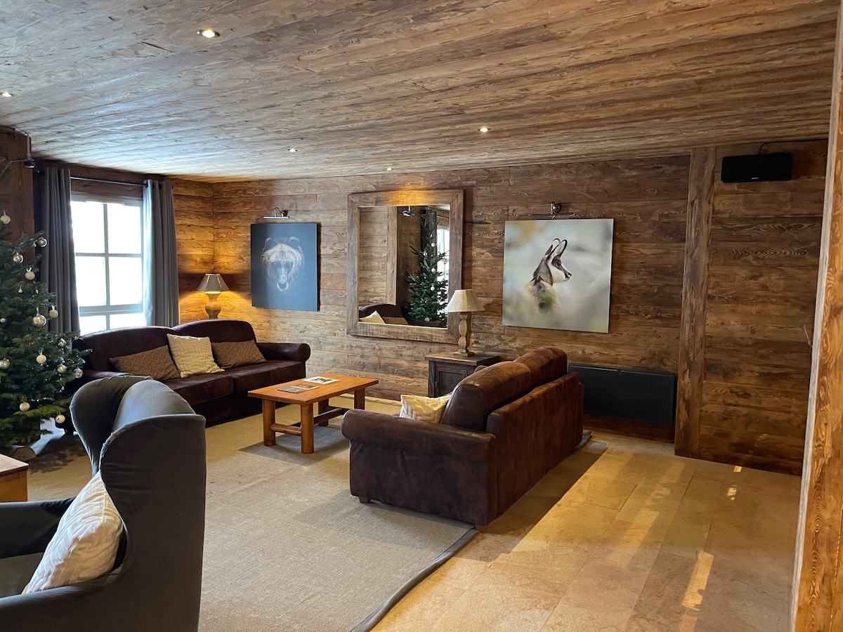 A cozy living area features wooden walls and a warm ambiance. Two brown sofas face a wooden coffee table. Art pieces adorn the walls, reflecting a modern touch. A mirror adds depth, while a soft area rug complements the space. Natural light filters through window treatments.