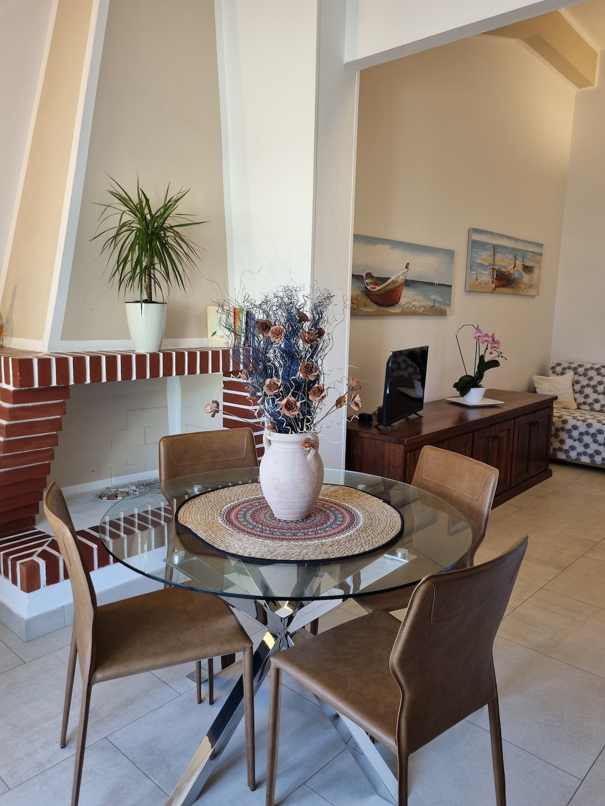 A modern dining area features a glass table surrounded by four brown chairs. A vase with decorative branches and dried flowers sits at the center. A fireplace with a plant above it and artwork depicting boats adorn the walls, enhancing the inviting atmosphere.