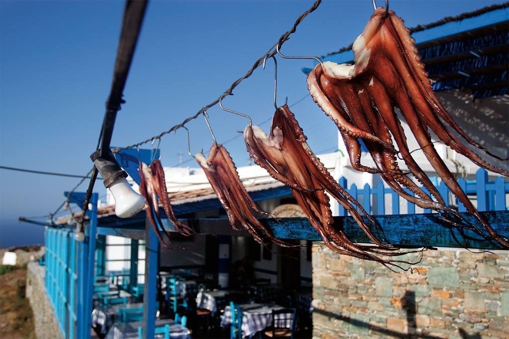 Dried octopuses are suspended on a line, displaying their rich, reddish-brown hues. A rustic blue and stone structure with tables is visible in the background, set against a clear blue sky. The scene offers a glimpse into local culinary traditions.