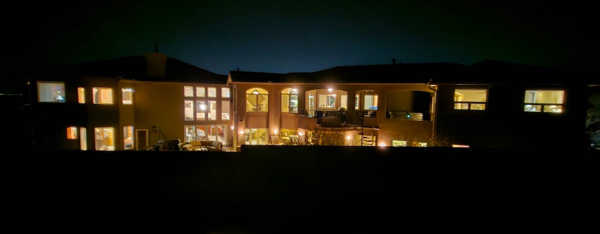 The exterior of a spacious home is illuminated at night, showcasing multiple large windows and balconies. Soft lighting defines various areas, highlighting outdoor seating and greenery. The structure features a warm color palette, contrasting beautifully against the dark night sky.