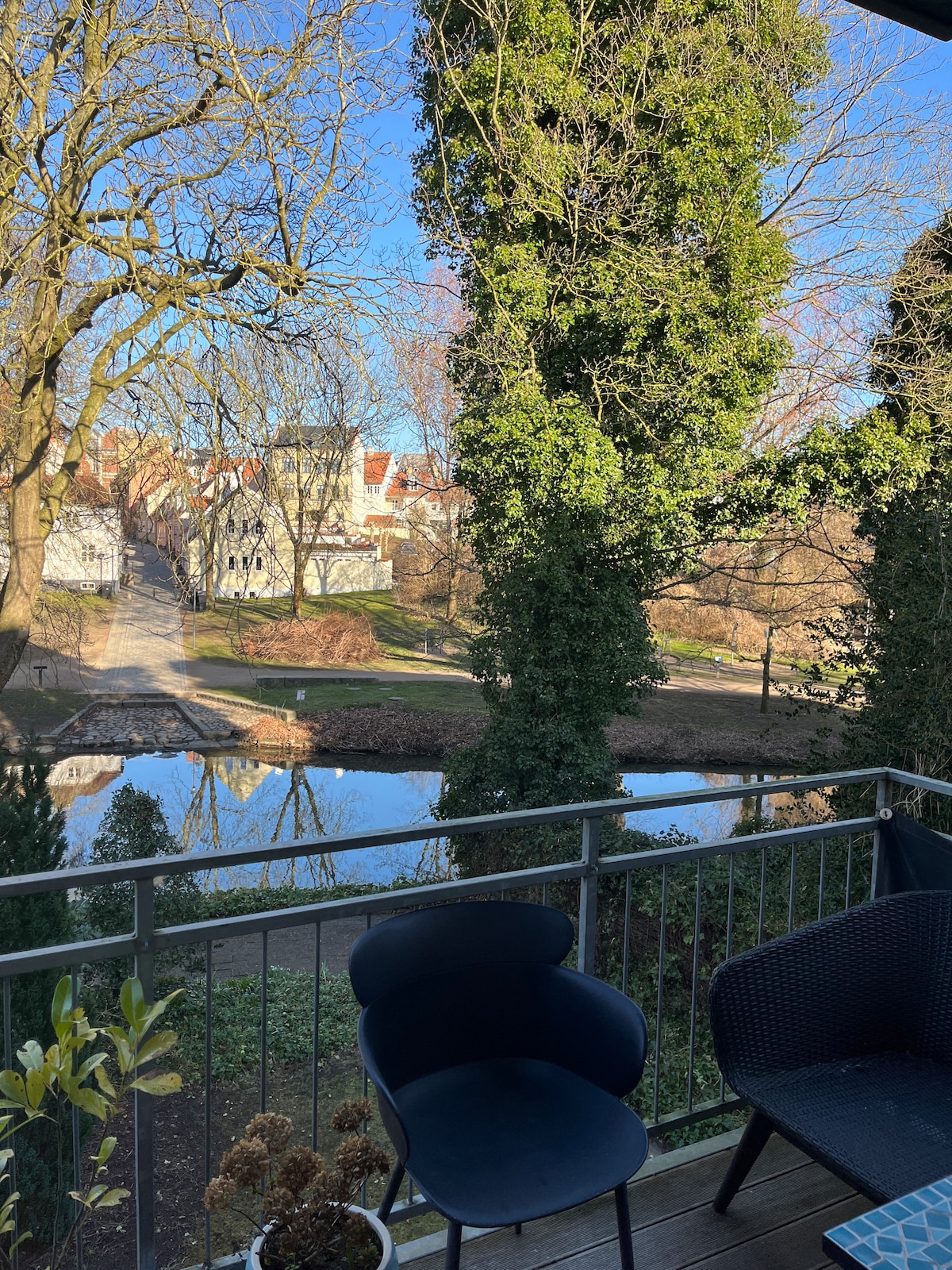 A serene balcony view showcases a tranquil waterway lined with trees and residential buildings in the distance. Two chairs sit on the balcony, surrounded by greenery, while sunlight filters through the branches, creating a peaceful outdoor setting.