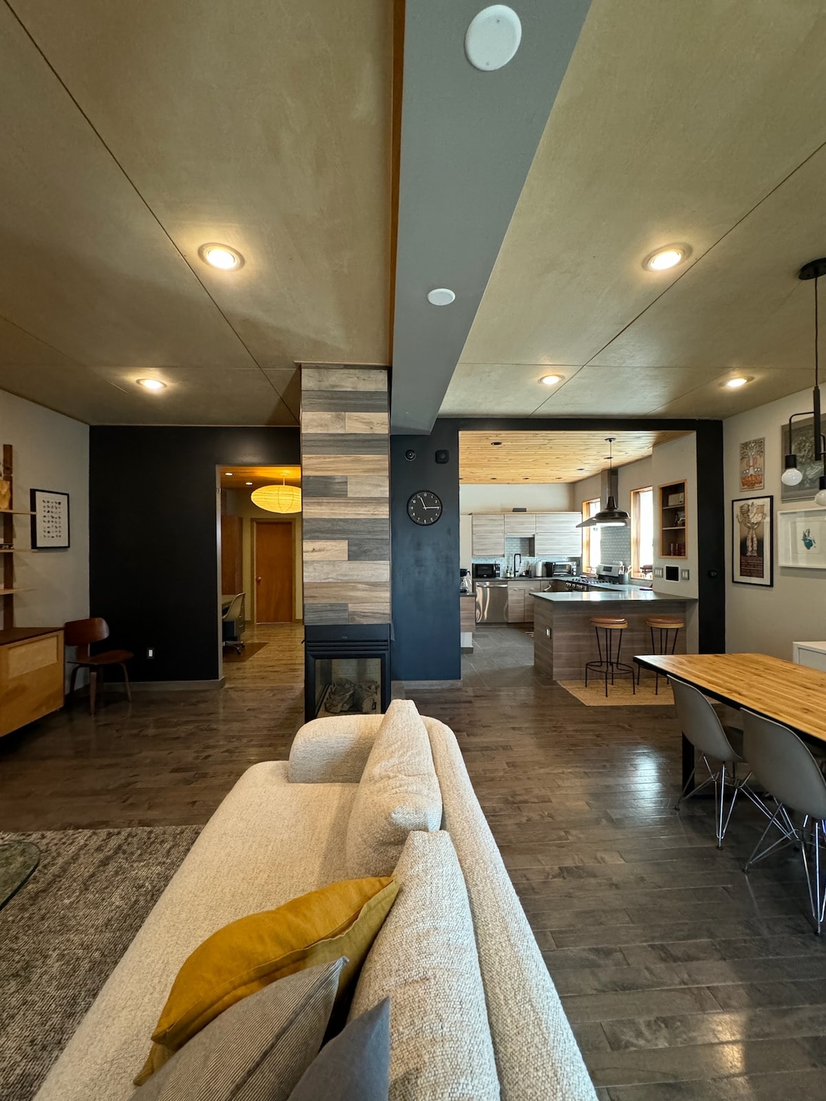 An open living space features a light-colored couch with yellow accent pillows. A wood accent wall adds texture, while recessed lighting illuminates the area. A dining table with chairs is visible in the background, leading towards a modern kitchen with an inviting atmosphere.