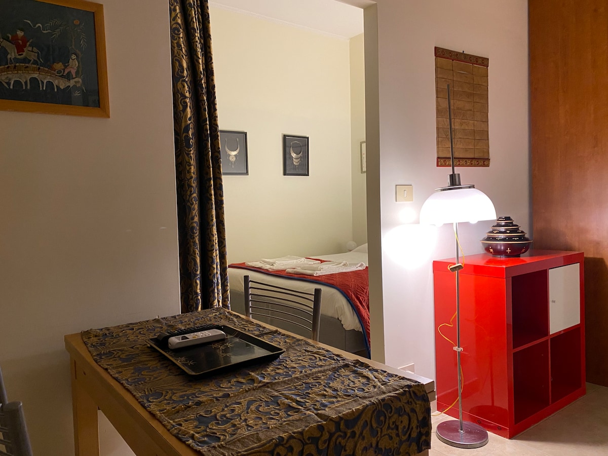 A warm and inviting living space is depicted, featuring a small dining table set with a tray and remote. A cozy bedroom is visible in the background, showcasing a neatly made bed with white linens. A soft glow is provided by a modern lamp beside a red shelving unit.