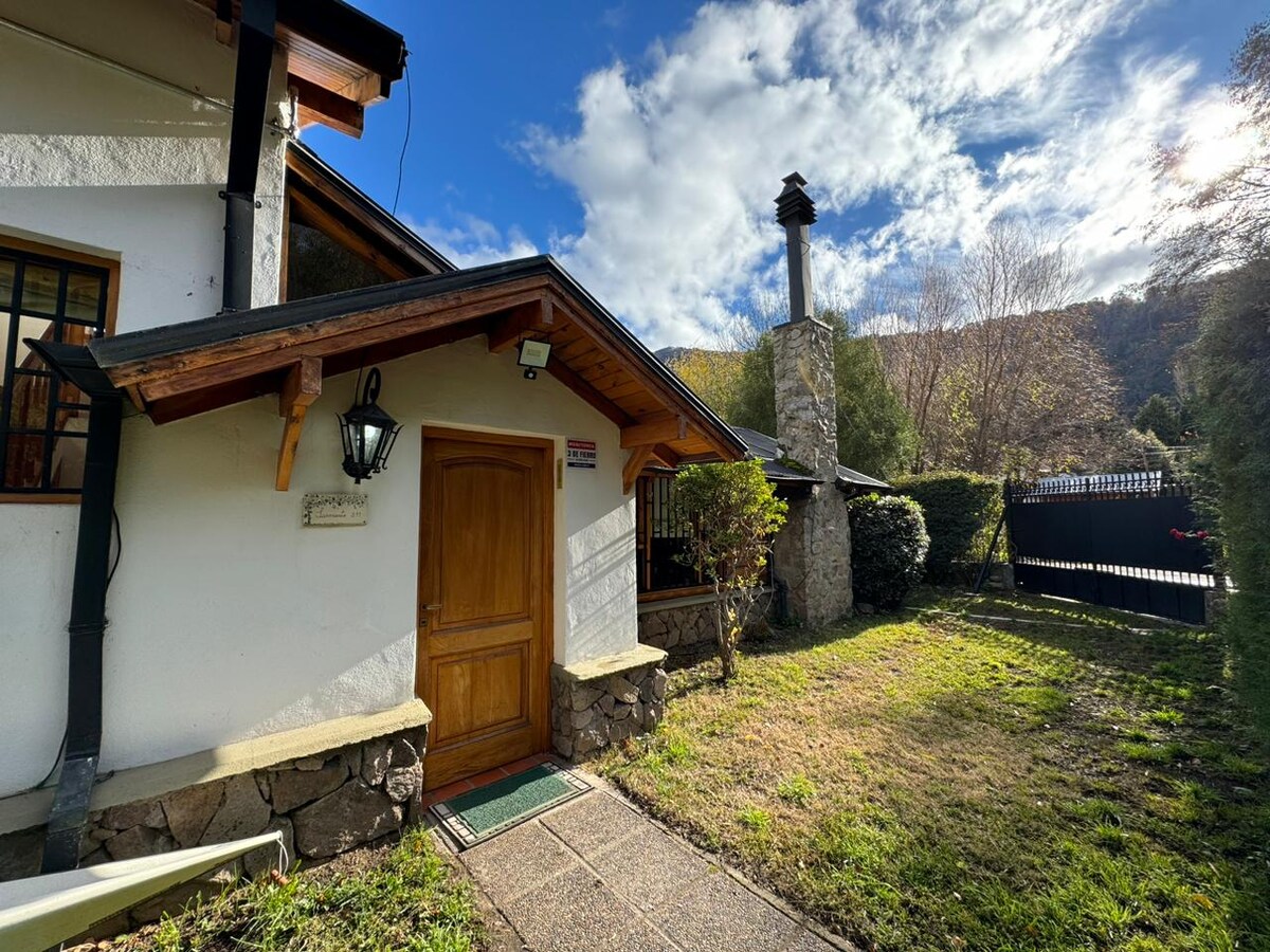 The entrance features a wooden door framed by stone and wood details. A lamp is mounted beside the door. A well-maintained garden with grass and shrubs surrounds the entryway, and a stone chimney is visible in the background, with a wooden fence marking the property boundary.