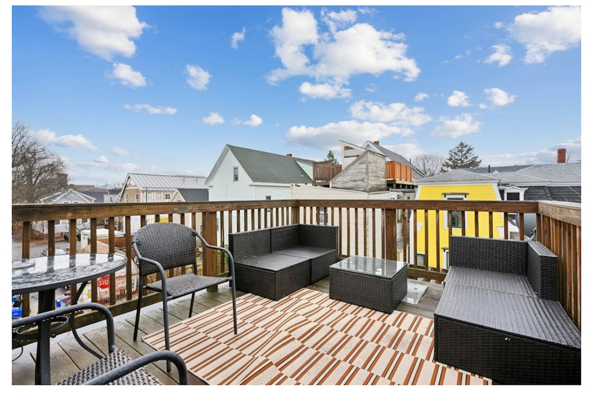 A spacious outdoor deck is featured, furnished with a seating area comprising a sectional sofa, coffee table, and dining table. A striped area rug adds warmth to the space. Clear skies and a view of neighboring rooftops create an inviting environment for relaxation.