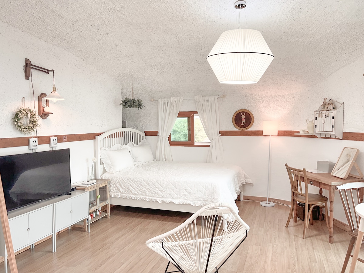 A bright and airy room features a spacious bed with white linens, complemented by soft lighting from a modern pendant lamp. A small dining table and chairs are placed alongside a minimalist TV unit, with a window providing a view of the surrounding greenery.