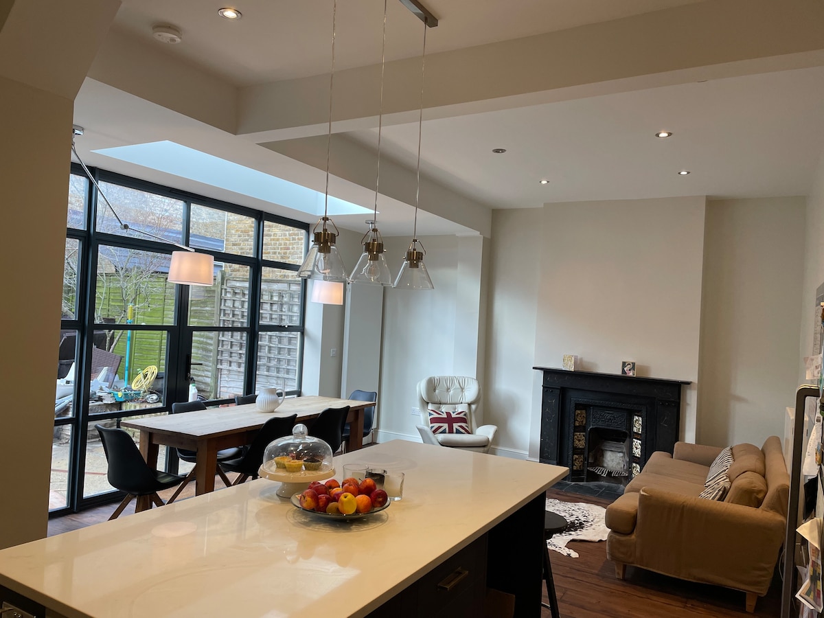 A modern kitchen diner is highlighted with a large island and pendant lighting. Natural light floods the open space through expansive glass doors leading to a garden. A dining table and additional seating area, featuring a fireplace, offer a welcoming atmosphere for family gatherings.