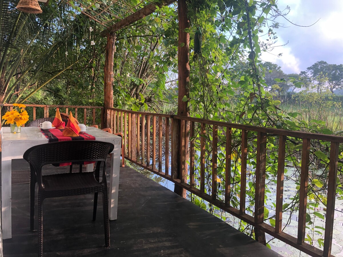 An outdoor dining area is surrounded by lush greenery, featuring a wooden railing that offers views of the serene lake. A white table is set with colorful napkins and a vase of flowers, enhancing the tranquil setting.