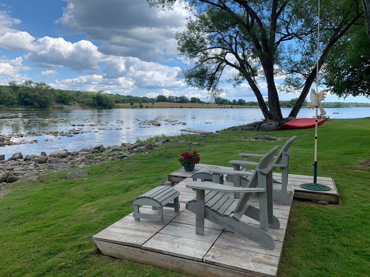 A scenic view of the river is framed by lush green grass and a majestic willow tree. Adirondack chairs are positioned on a wooden deck, with a small table beside them. A kayak hangs nearby, suggesting outdoor activity by the water's edge.
