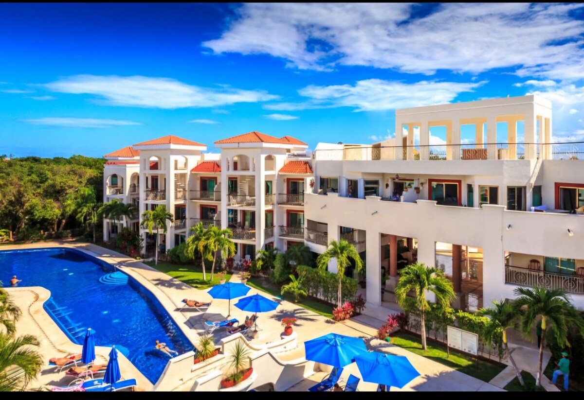 A landscaped area showcases a large pool surrounded by sun loungers and umbrellas. The multi-level condominium building features multiple balconies and terraces, with lush greenery enhancing the tranquil setting beneath a bright blue sky.