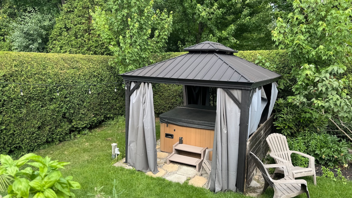 A private hot tub is situated under a gazebo, surrounded by lush greenery and hedges. Light grey drapes frame the opening of the gazebo, providing an inviting atmosphere. Comfortable adirondack chairs are positioned nearby on a stone patio, complementing the tranquil outdoor setting.