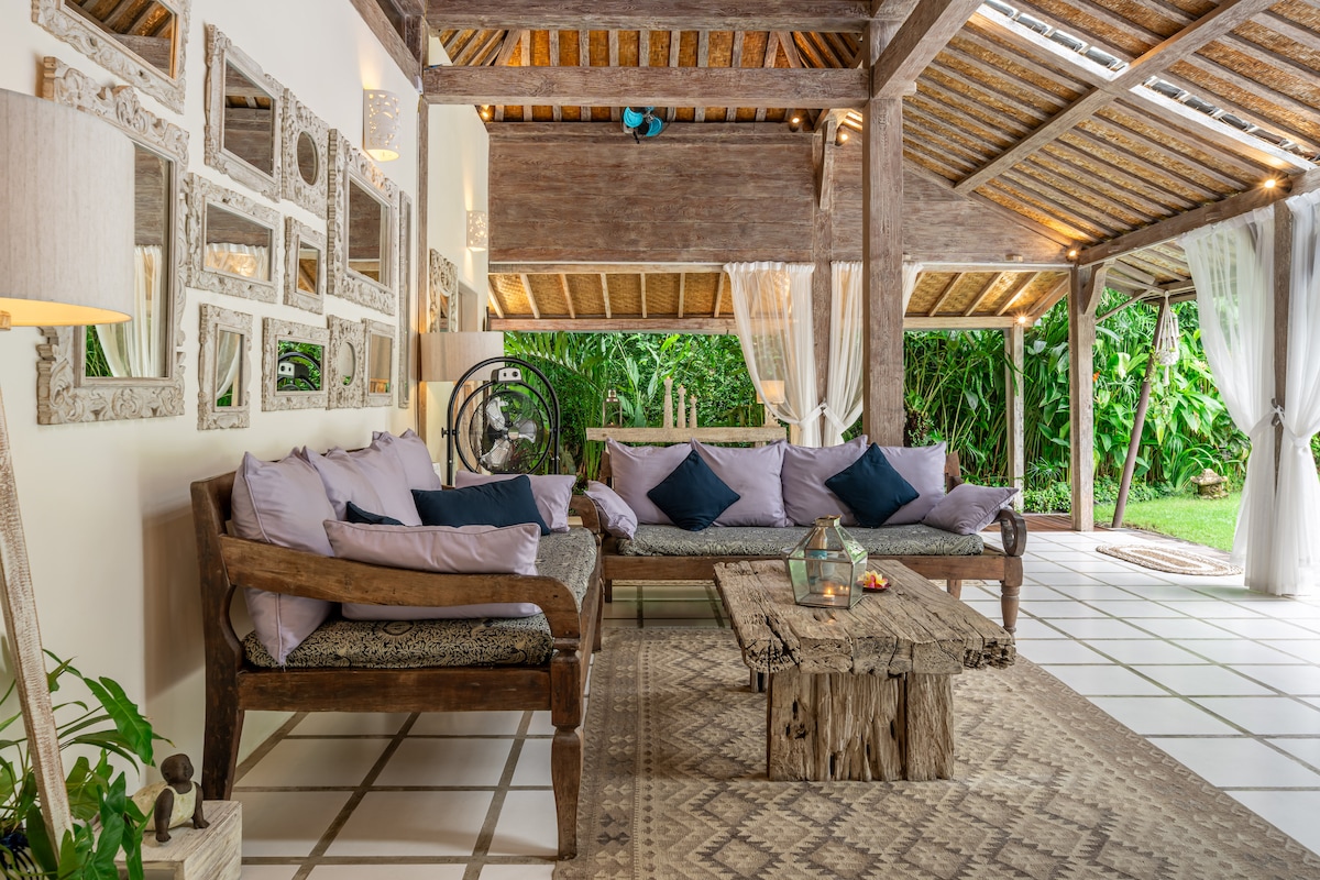 An open living area features wooden beams and a high ceiling, complemented by a mix of natural materials. Soft seating with purple cushions surrounds a rustic wooden coffee table, while mirrors adorn the wall. Lush greenery is visible through the glass doors, enhancing the space.