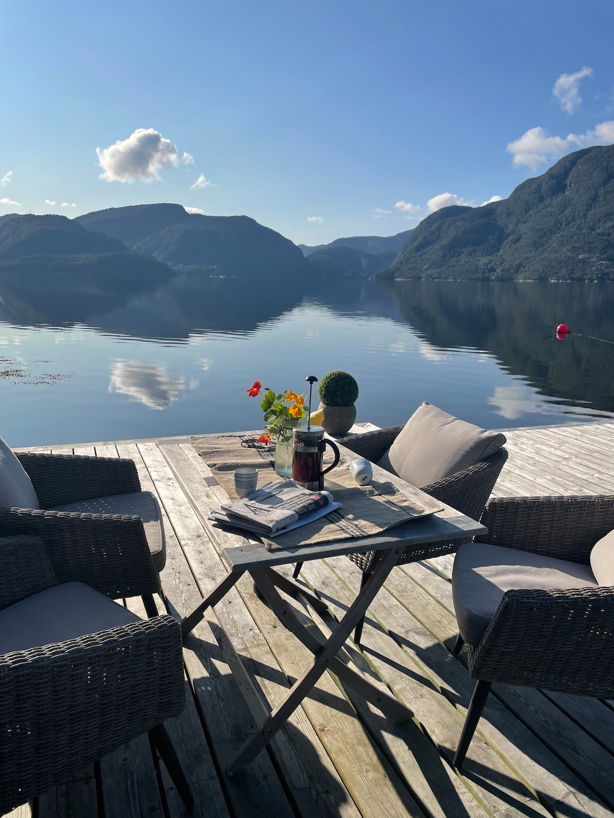 A table is set on a wooden deck overlooking calm waters, surrounded by mountains. Two comfortable chairs frame the table, which holds a vase of flowers and a coffee pot. Soft sunlight enhances the serene environment, reflecting off the water's surface.