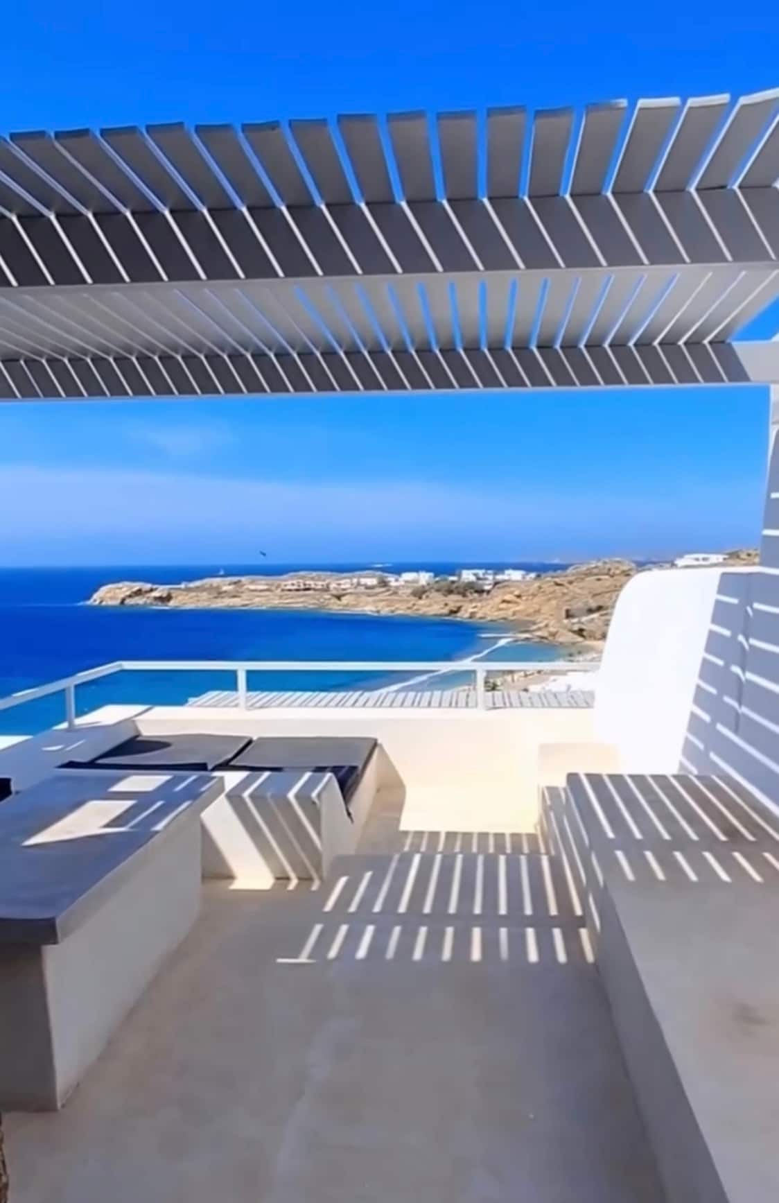 An outdoor terrace showcases a dining area with a table and seating, framed by a pergola. A panoramic sea view extends beyond, featuring clear blue waters and rocky coastline under a bright sky. Shadows cast by the pergola create a striped light pattern on the floor.