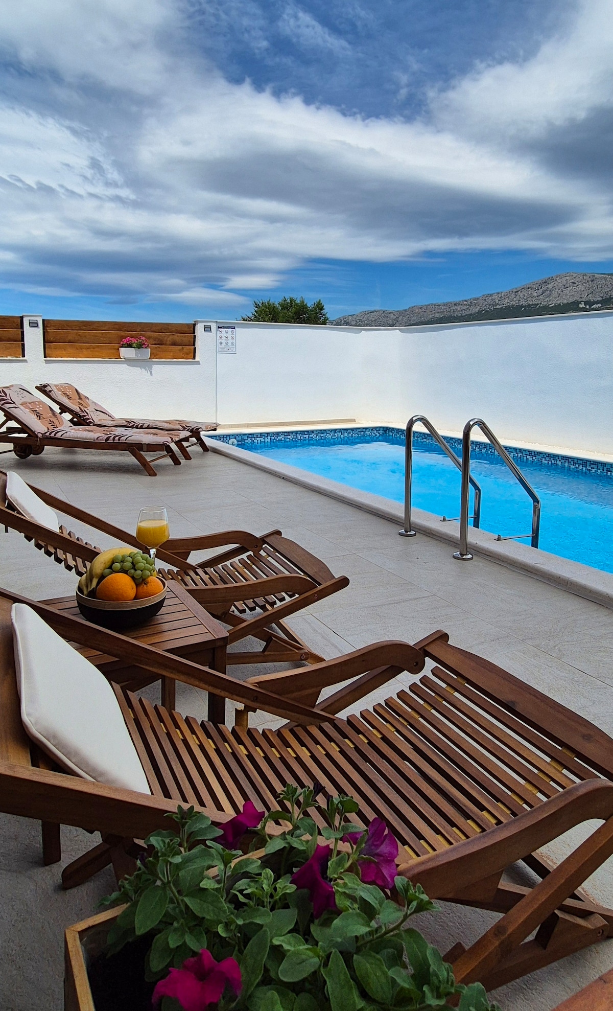 A private outdoor swimming pool is set against a clear blue sky, with sun loungers arranged nearby. A bowl of fresh fruit and a glass are positioned on a table, while vibrant flowers add color to the scene, inviting relaxation.