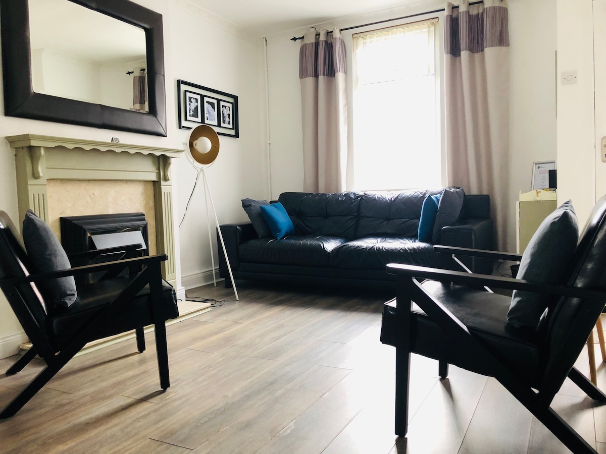 A spacious living area is highlighted by a large black leather sofa and two black armchairs, arranged around a light wooden floor. A traditional fireplace is visible with a large mirror above it, and a window with light curtains provides natural light.