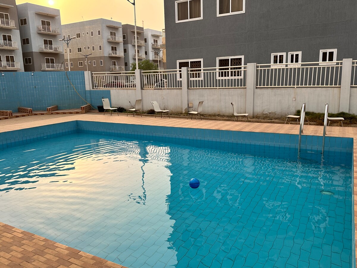 A community swimming pool is featured, reflecting the warm hues of the setting sun. The pool area includes a shallow end with swimming steps and a floating ball. Sun loungers are positioned along the edge, while surrounding buildings are visible in the background.