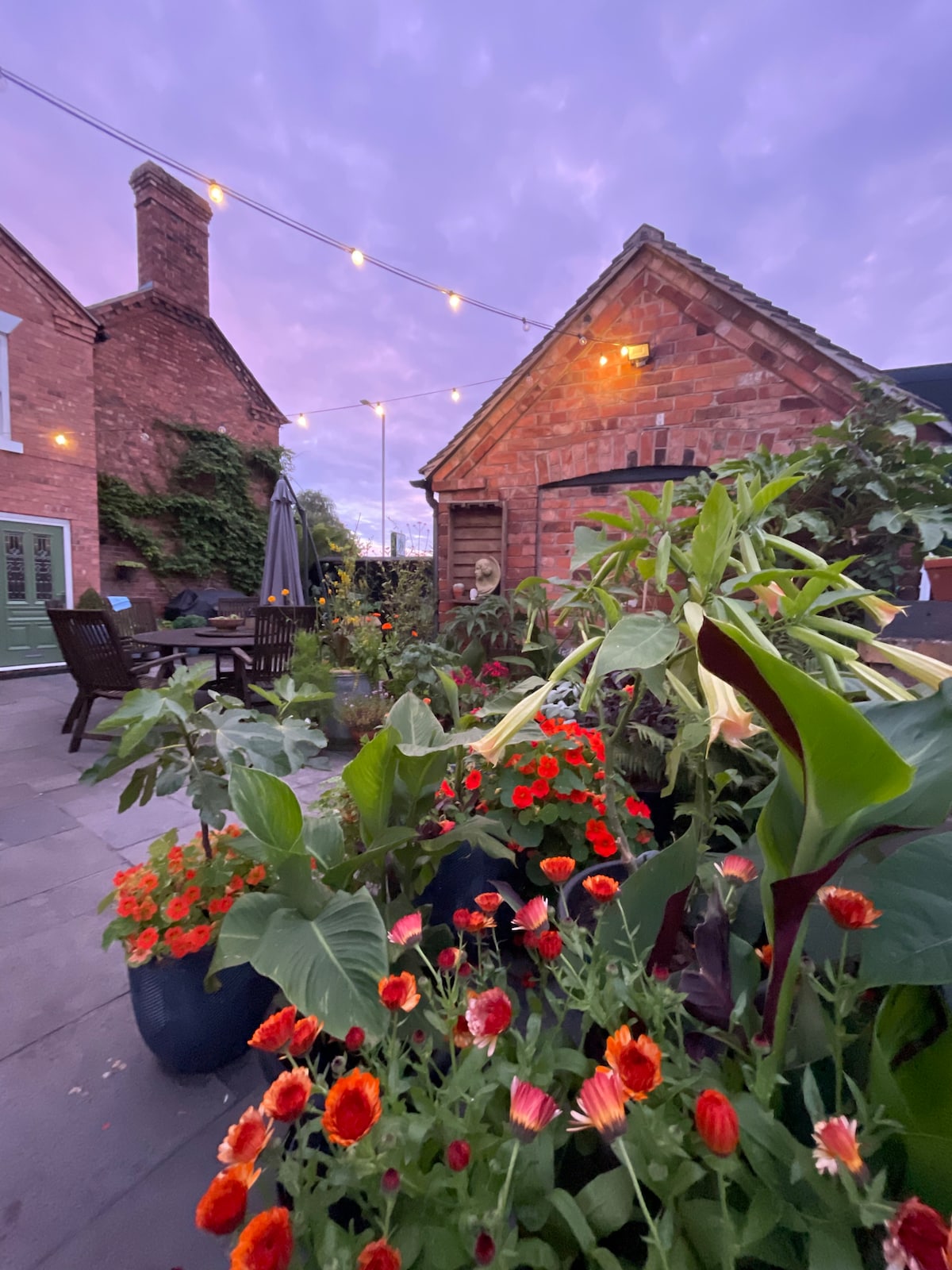 A vibrant courtyard garden is featured, adorned with a variety of lush flowers in bloom. Garden furniture is visible among the plants, with warm lighting illuminating the area at dusk. The rustic brick buildings provide a charming backdrop to the greenery.