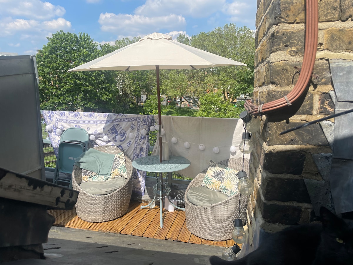 A private outdoor terrace is depicted, featuring a round table and two comfortable chairs. A large umbrella provides shade, while decorative lights are hung nearby. Lush greenery is visible in the background, enhancing the serene atmosphere.
