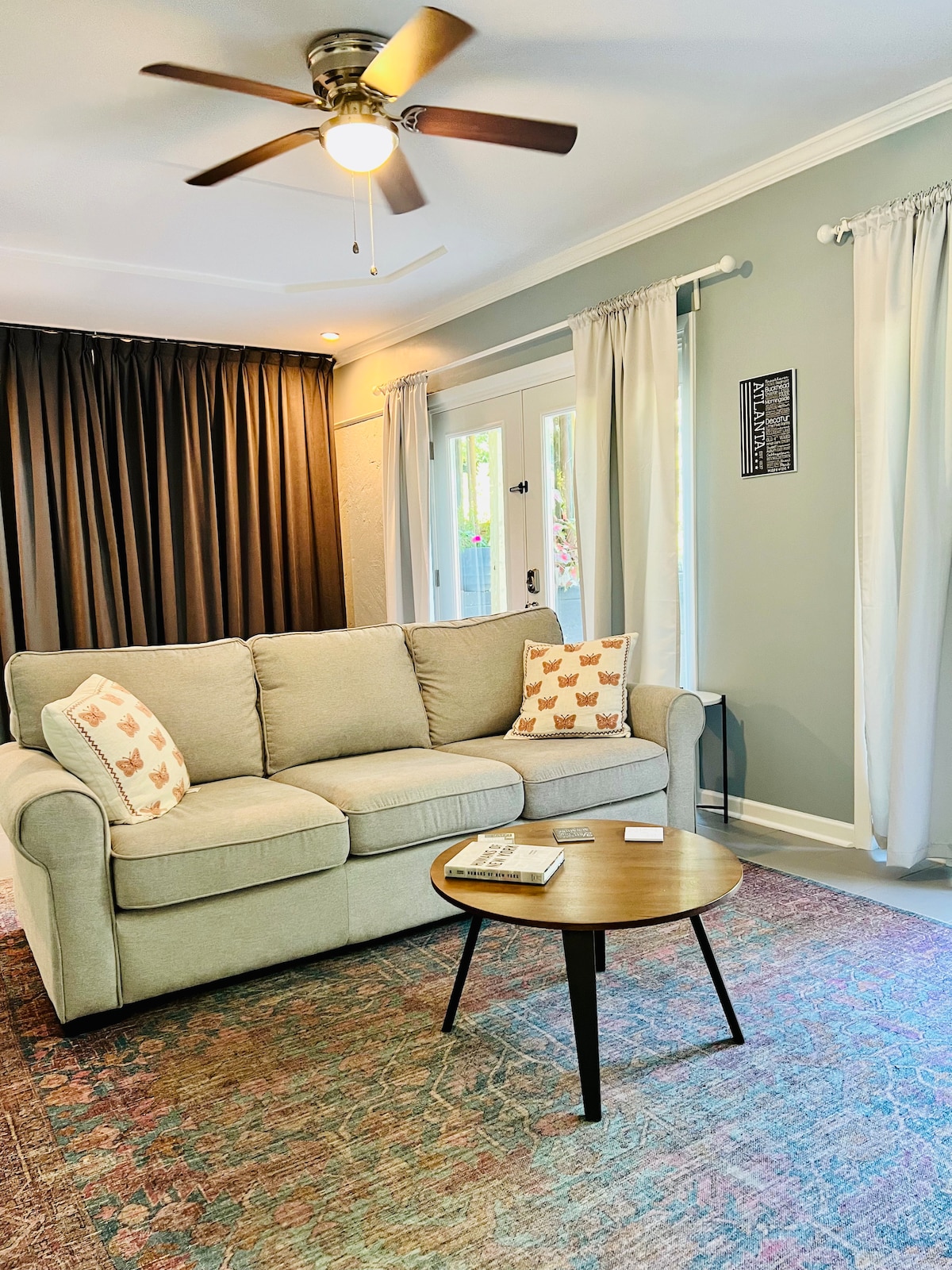 A comfortable living area features a light-colored sofa adorned with patterned cushions. A round coffee table is placed in front, accompanied by a decorative rug. Natural light filters through sheer white curtains, while dark curtains add depth to the space.