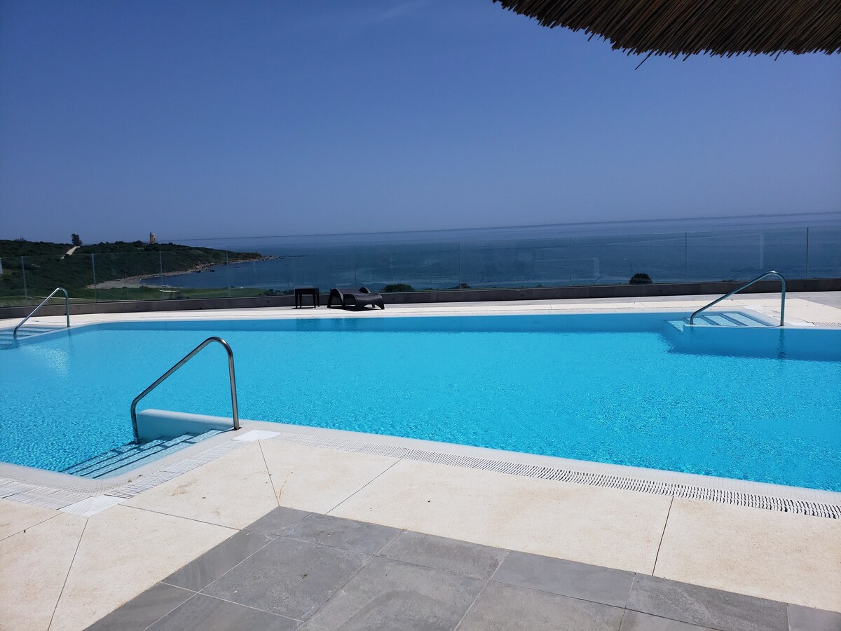 An outdoor pool is framed by a wide deck, featuring steps leading down into the water. The pool’s vibrant blue hue reflects the clear sky and ocean beyond, offering expansive views of the coastline. Loungers are positioned nearby for relaxation.