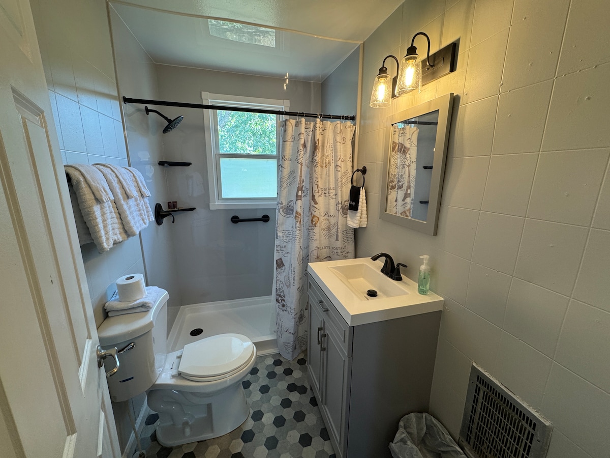 A small full bath features a modern vanity with a sink and a mirror above. White and gray hexagonal tile covers the floor. A white bathtub with a shower curtain was shown, along with neatly arranged towels on the towel rack. Natural light filters in through a window.