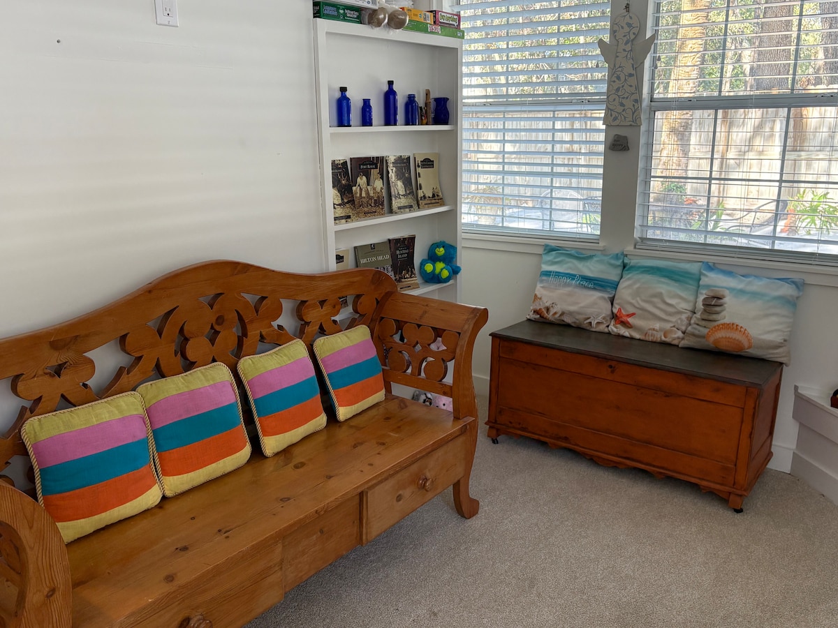 A cozy sunroom features a wooden bench with colorful striped cushions, inviting relaxation. A rustic wooden chest sits nearby. Natural light filters through the partially open blinds, illuminating shelves lined with books and blue decorative bottles.