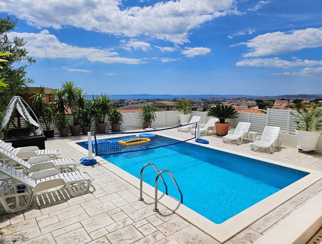 A private pool is featured in a sunlit area surrounded by tropical plants and plush loungers. The pool is equipped with a net for casual games. Scenic views of the landscape and city are visible beyond the pool, complemented by a clear blue sky.