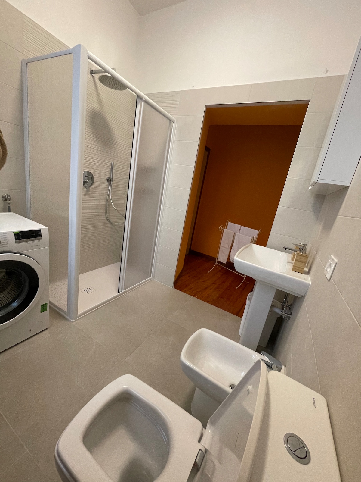 The bathroom features a walk-in shower enclosed by glass, coupled with a modern sink and vanity. A washing machine is positioned conveniently beside the shower. Two toilets are located in the space, which is adorned with neutral tiles and complemented by warm-toned wall paint.