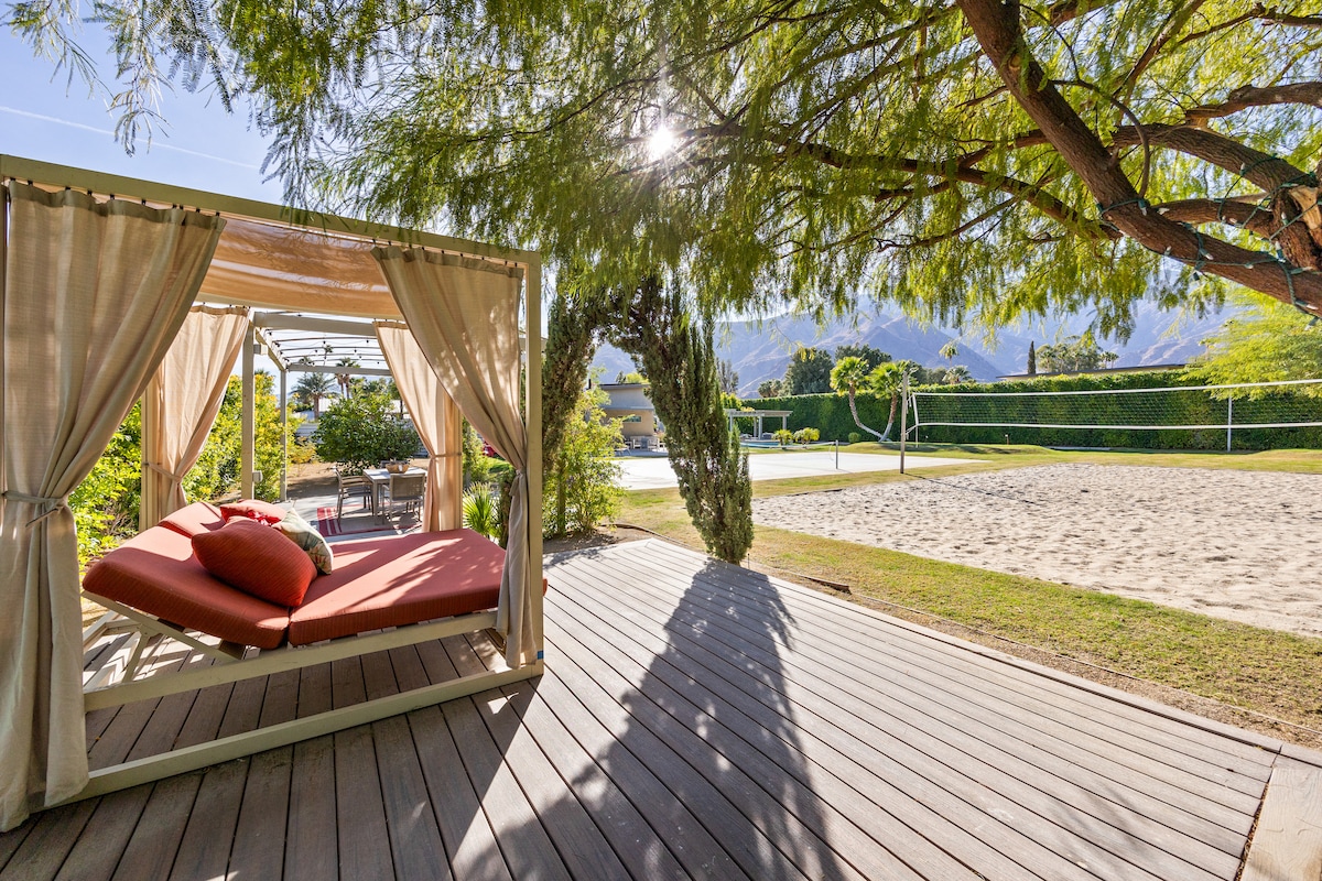 A shaded outdoor lounging area features a comfortable daybed with cushions, situated on a wooden deck. In the background, a sand volleyball court is visible alongside lush green lawn areas and distant views of the surrounding mountains under clear skies.